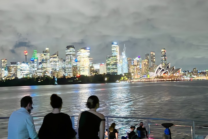 Three people sit by a waterfront with city skyline and opera house in the background at night.