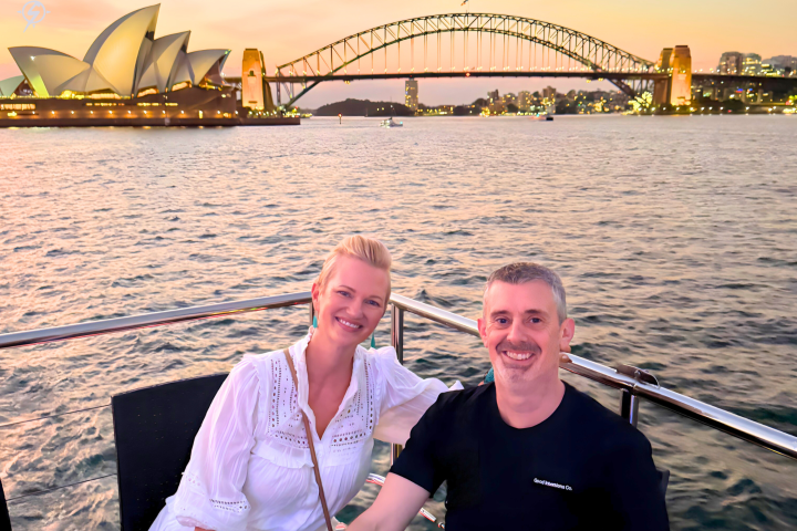 Two people smiling on a boat with Sydney Opera House and Harbour Bridge at sunset.