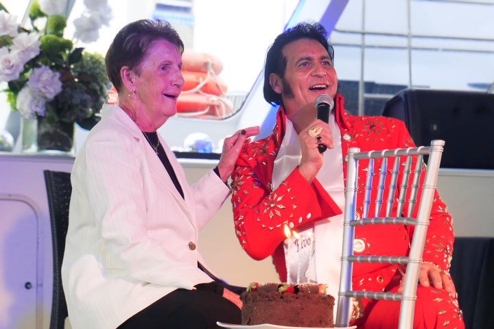 Woman and man in red suit with microphone share a moment near a cake on a boat.