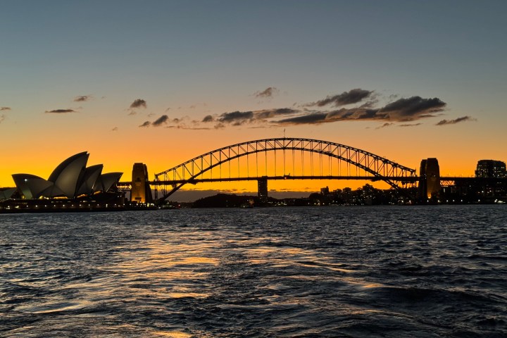 Sydney Opera House and Harbour Bridge at sunset with water in foreground.