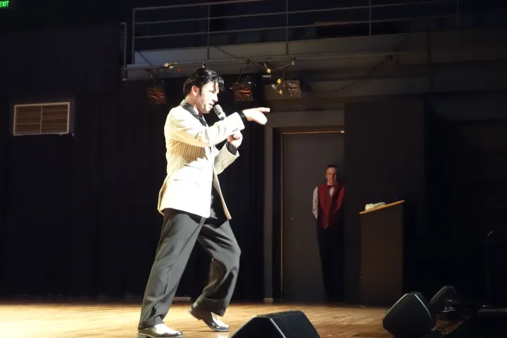 Performer on stage singing into a microphone, person in red vest watching from doorway.