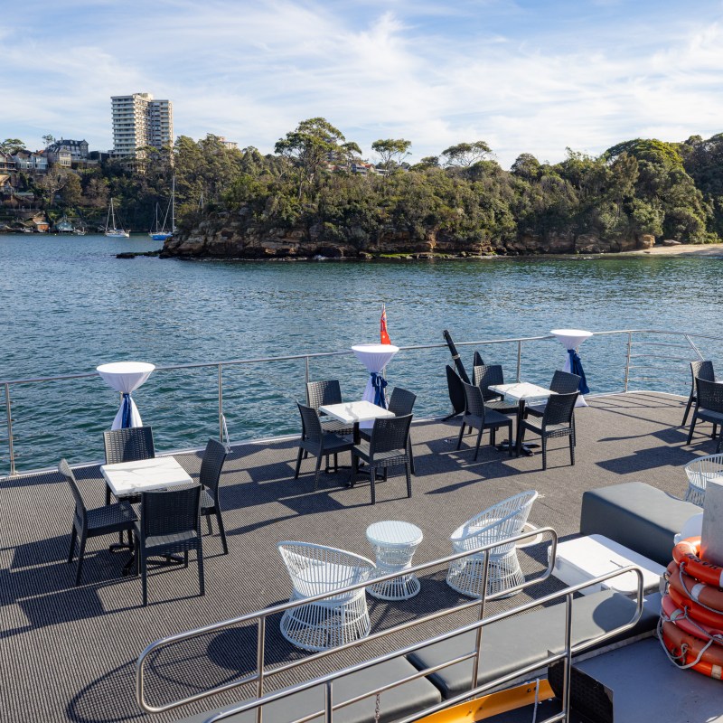 Outdoor deck of a boat with tables and chairs overlooking a bay and wooded shoreline.