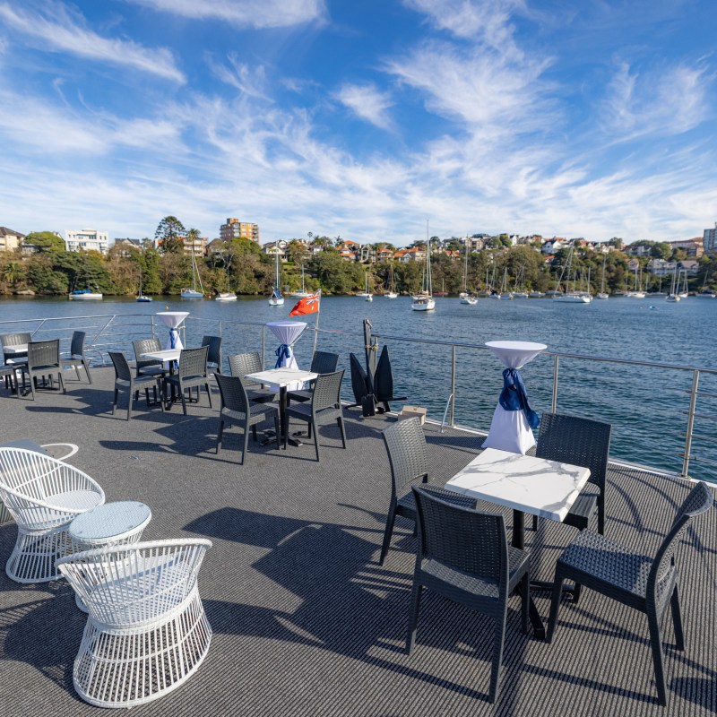 Boat deck setup with tables and chairs, overlooking water with distant sailboats and city skyline.