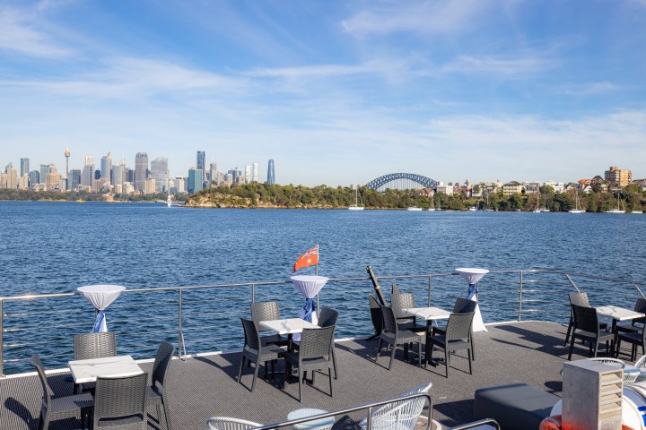 Boat deck with tables and chairs, overlooking a city skyline and bridge.