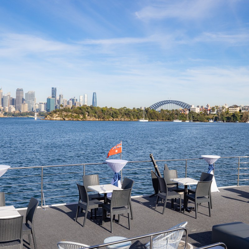 Boat deck with tables and chairs, overlooking a city skyline and bridge.