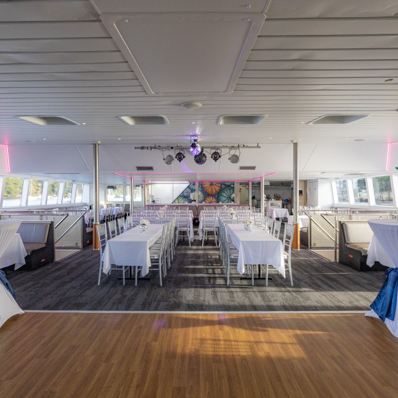 Interior of a decorated event space on a boat with tables and chairs, wooden flooring, and pink lighting.