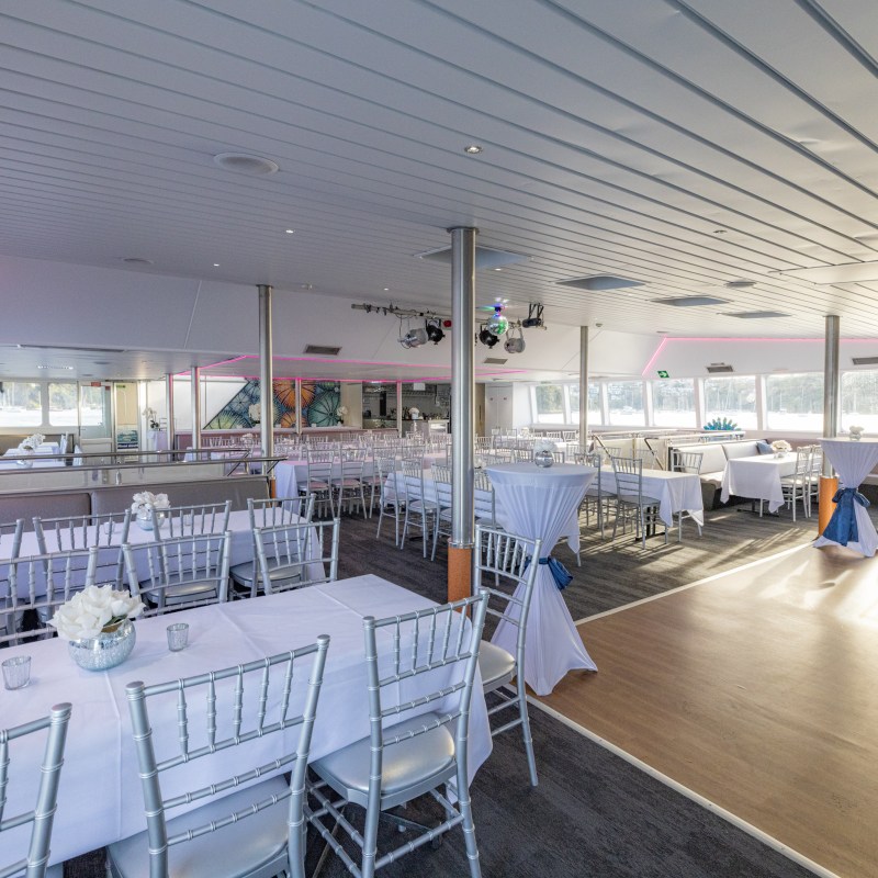 Spacious event space with white tables and chairs, pink lighting, and large windows.