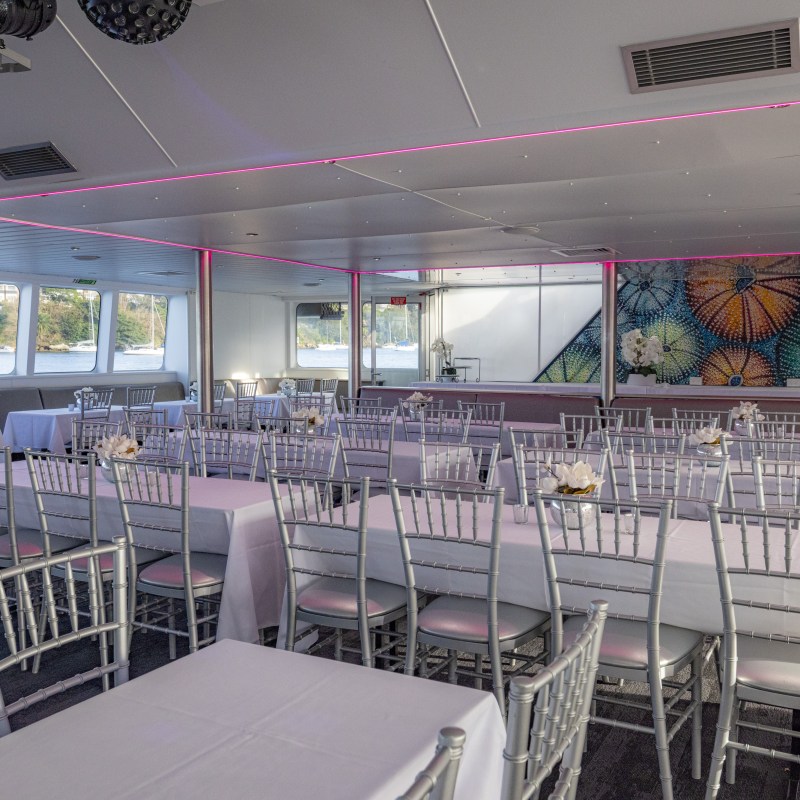 Event space with white tablecloths and silver chairs, featuring large windows and colorful wall art.
