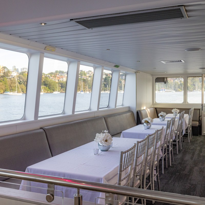 Interior of a boat with tables and chairs, windows showing water and shoreline outside.