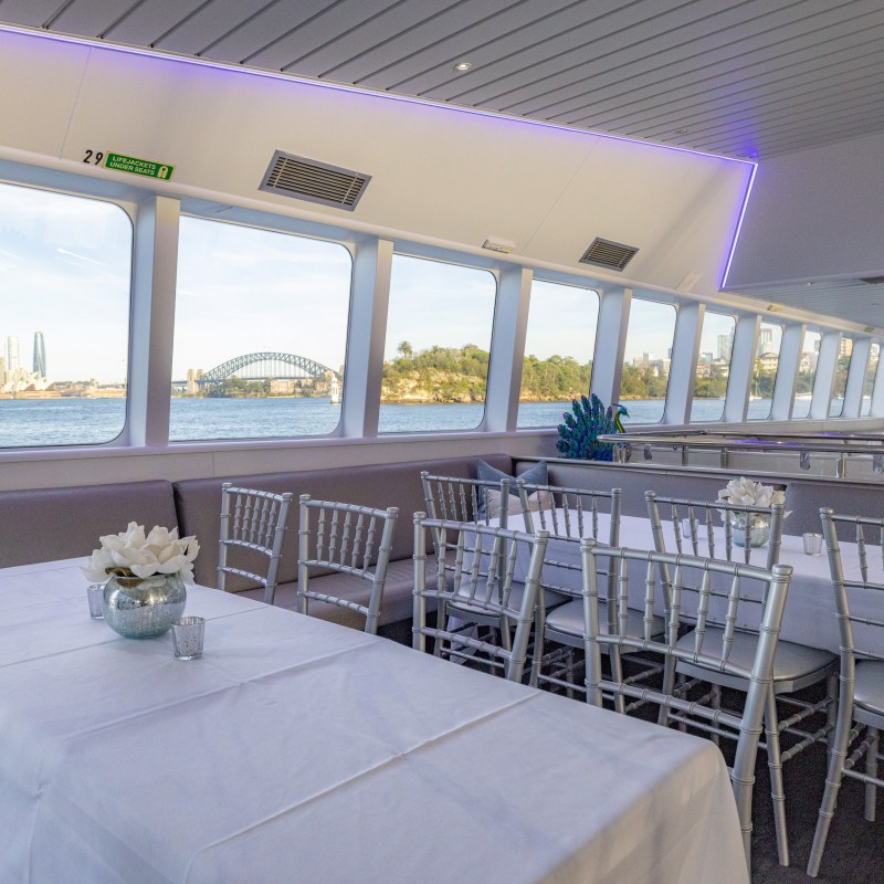 Interior of a boat with dining tables, chairs, and a view of a cityscape and bridge through large windows.