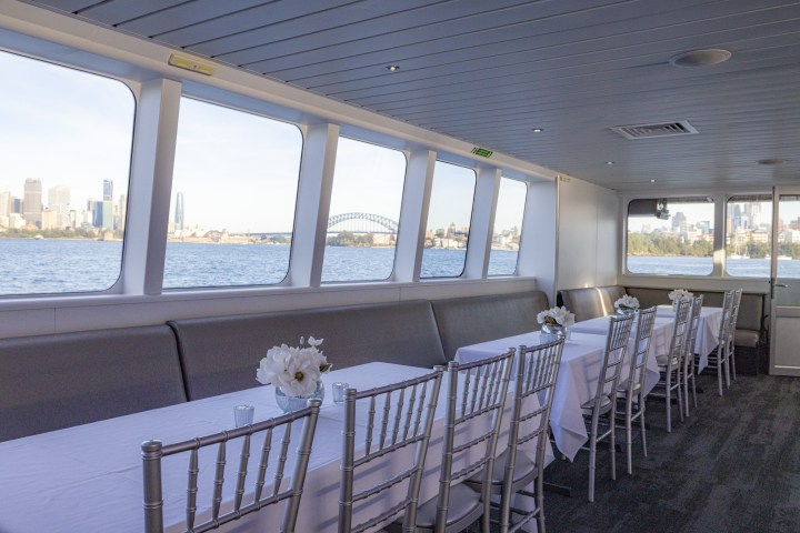 Interior of a boat with tables and chairs, large windows showing a city skyline and bridge over water.