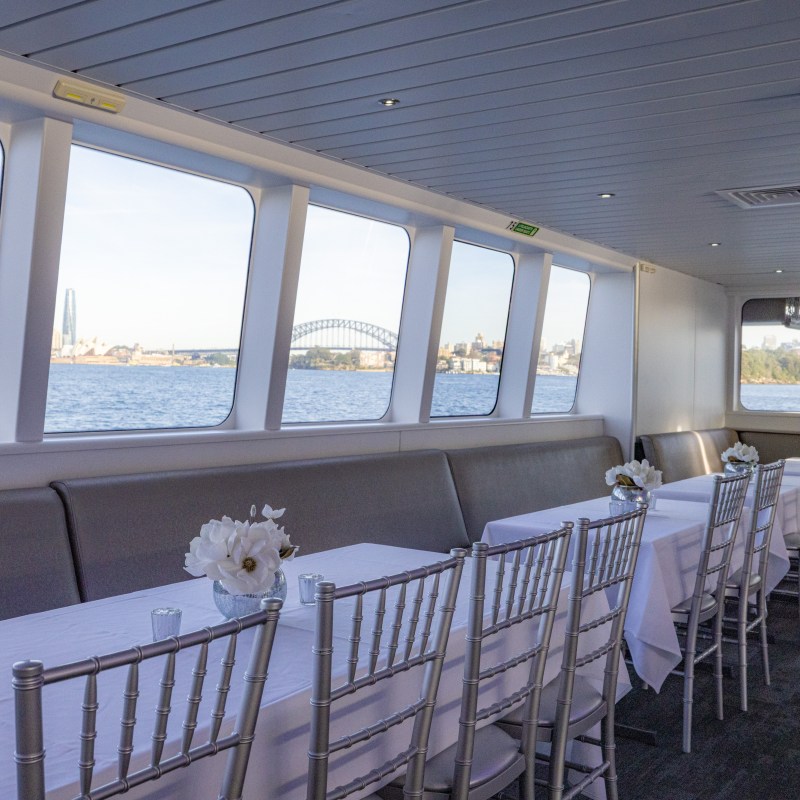 Interior of a boat with tables and chairs, large windows showing a city skyline and bridge over water.