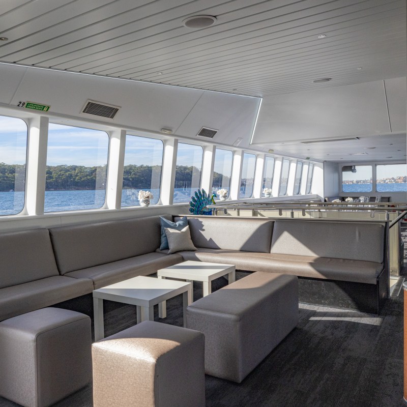 Modern boat interior with gray sofas, low tables, and large windows overlooking water and trees.