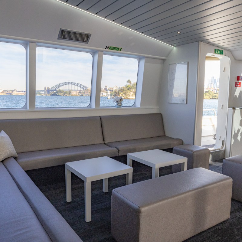 Interior of a boat lounge with gray seating, tables, and windows showing a cityscape with a bridge.