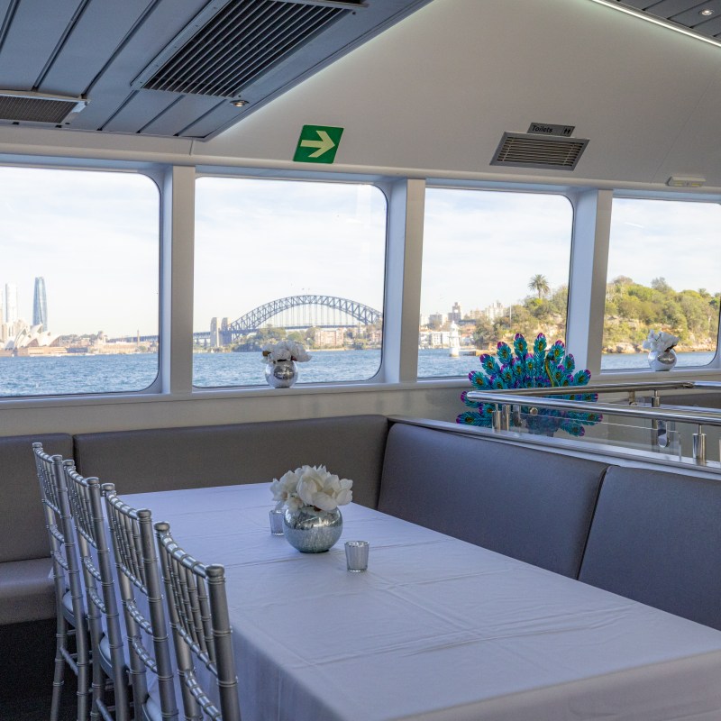 Interior of a boat with table and chairs, view of Sydney Opera House and Harbour Bridge through windows.