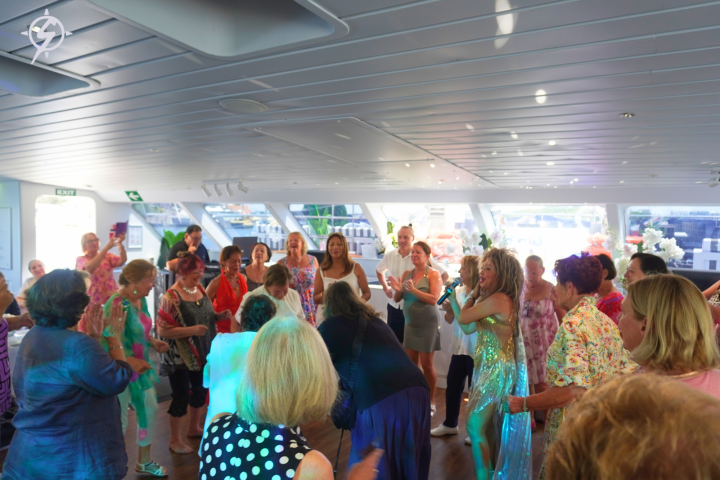 Group of people dancing indoors on a boat with a singer in a silver dress.
