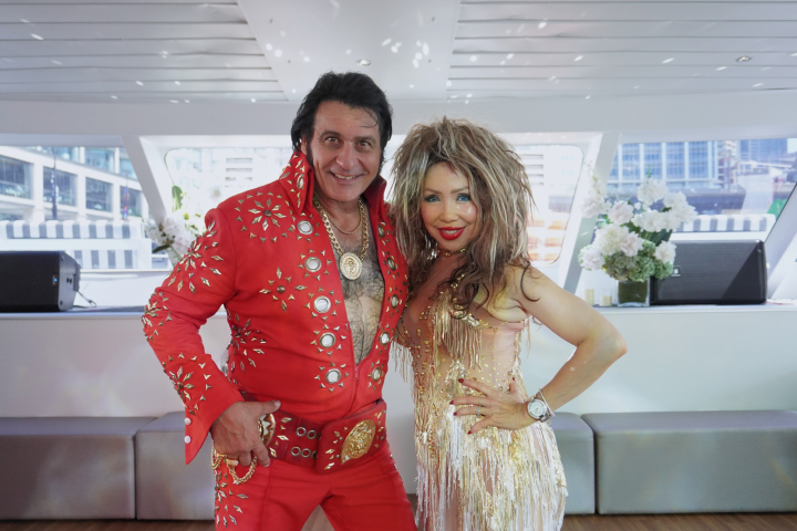 Two people in stage costumes with disco-themed decor, one in red and one in gold, posing together.