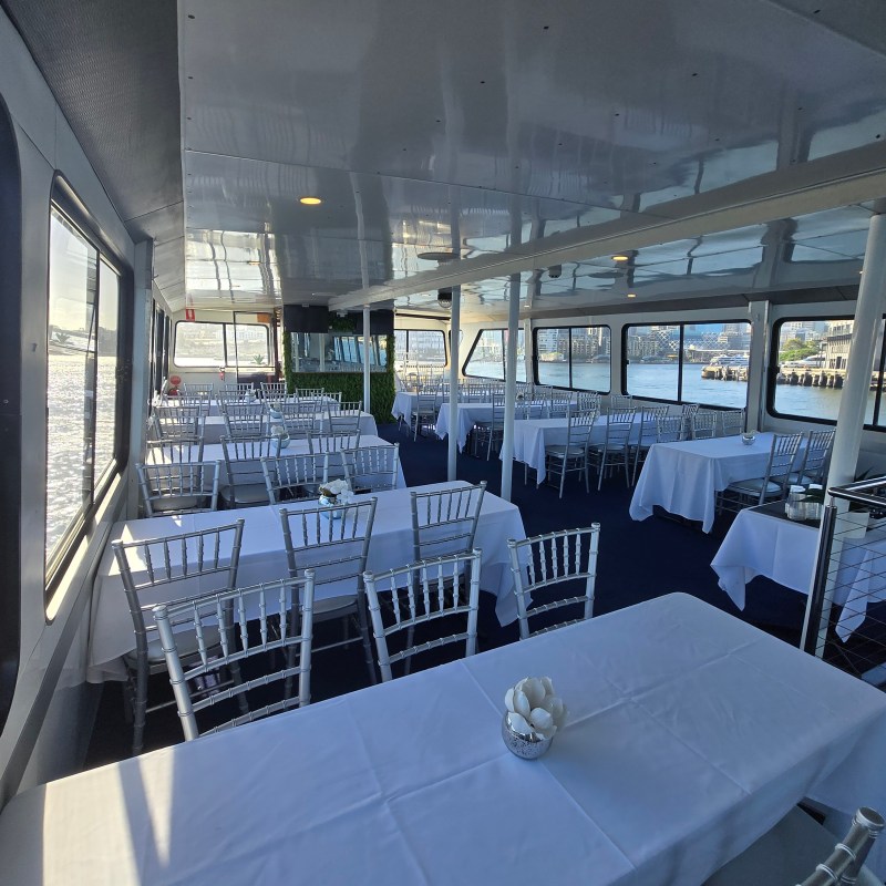 Interior of a boat with tables and chairs set for dining, positioned near large windows overlooking the water.