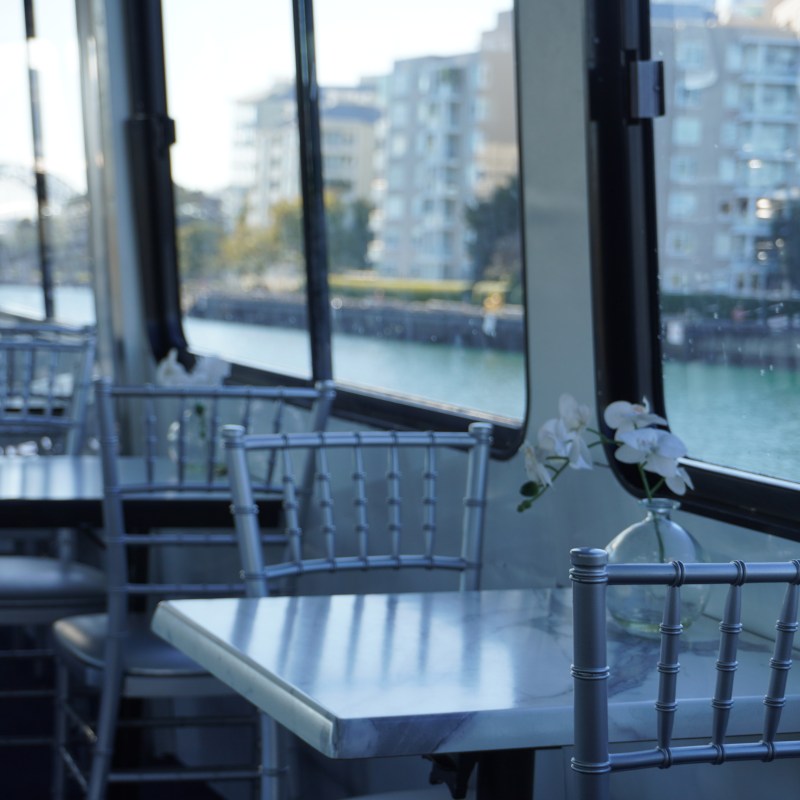 Empty ferry with window view of urban buildings over water, featuring tables and chairs.