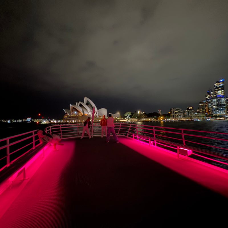 Dark sky over Sydney Opera House with pink-lit walkway and city skyline.